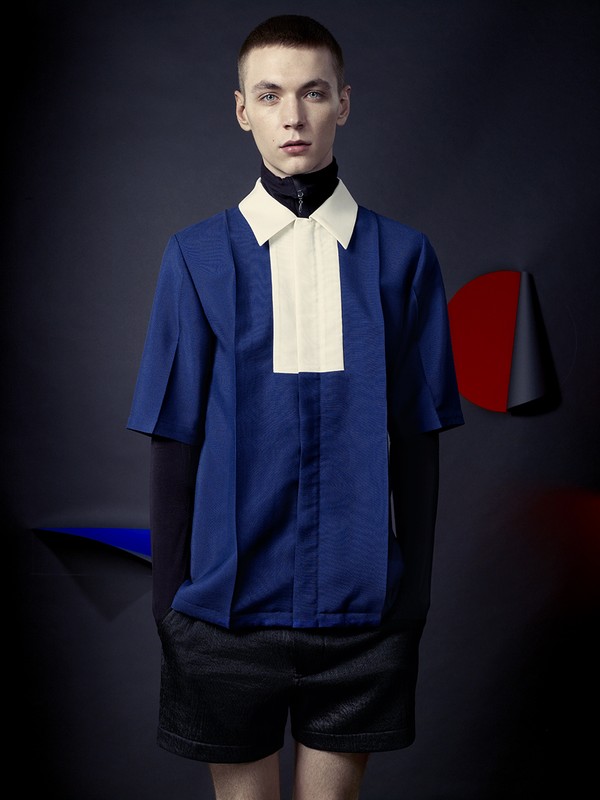 A fair-skinned young man wearing a blue short-sleeved shirt with a white collar and black shorts posing indoors with a dark background.