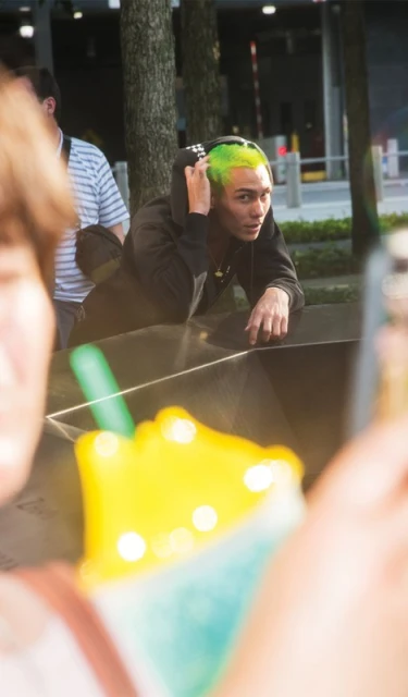 Young man with vibrant green hair and casual clothing, photographed outdoors with an out-of-focus beverage in the foreground.