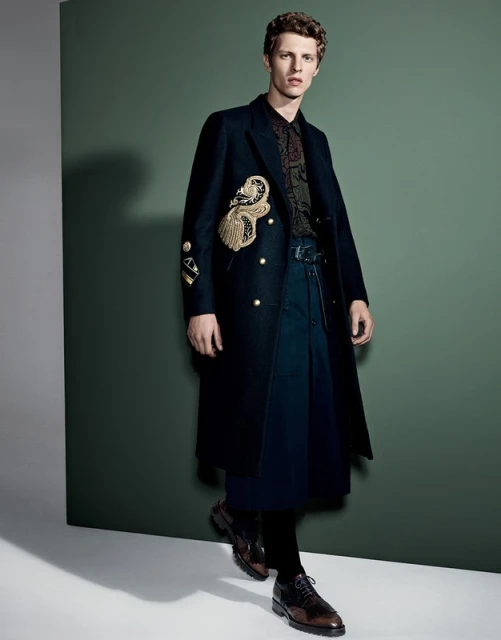 A curly-haired male model poses in a dark patterned shirt, blue bottoms, and a long black embellished coat against a green background.