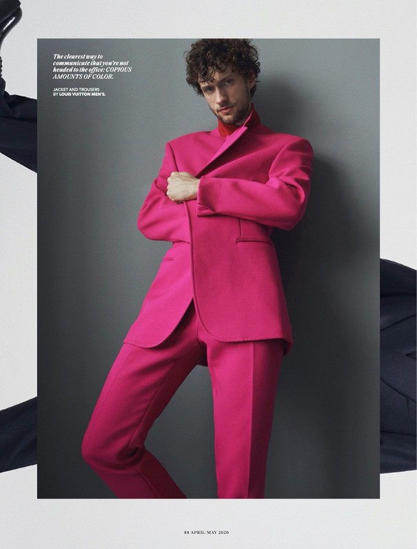 An adult male fashion model poses indoors, wearing a bright pink suit and a red undershirt, against a grey background in a fashion editorial.
