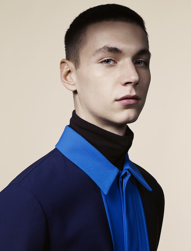 Portrait of a young adult man wearing a black turtleneck sweater and a blue collared shirt against a neutral background.