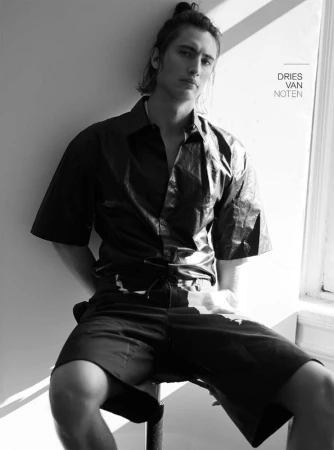 Monochrome portrait of a young male model with hair tied back, wearing a short-sleeve button-down shirt and shorts, seated indoors.