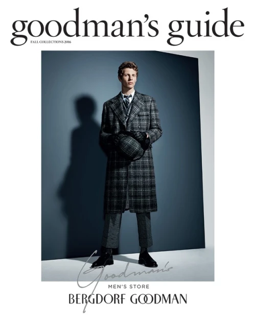 Full-length studio shot of a young adult male model wearing a grey plaid overcoat and pants with black boots against a grey backdrop.