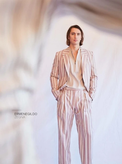 A fair-skinned adult male model poses indoors wearing a striped tan suit. The photograph features neutral colors and a blurred background.