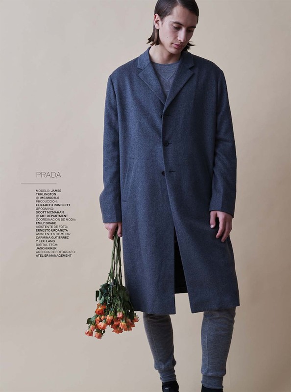 A young adult man in a grey coat holds orange roses against a cream backdrop for a fashion editorial photography shot.