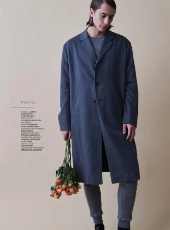 A young adult man in a grey coat holds orange roses against a cream backdrop for a fashion editorial photography shot.