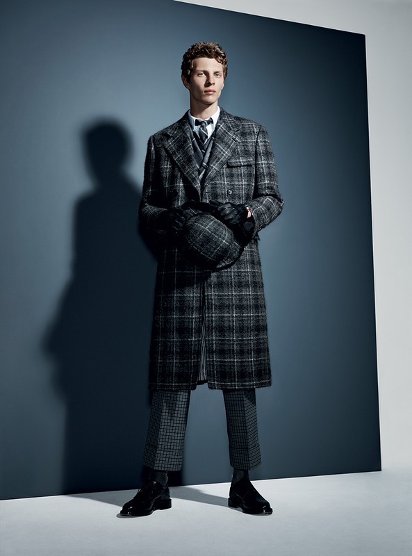 Man in a grey plaid coat and pants holding a hat, standing in a photo studio with a blue and grey background, fashion shot.