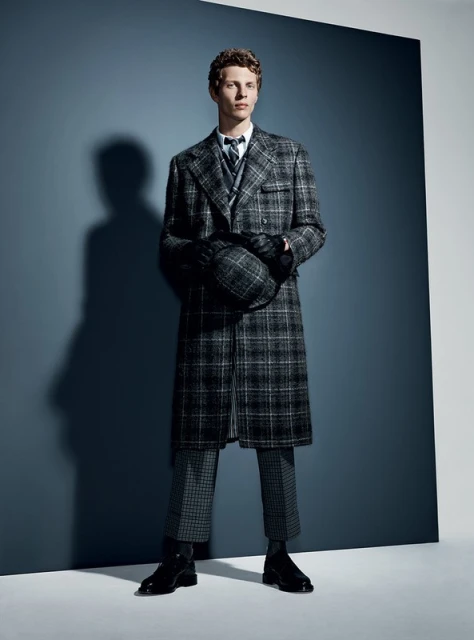 Man in a grey plaid coat and pants holding a hat, standing in a photo studio with a blue and grey background, fashion shot.