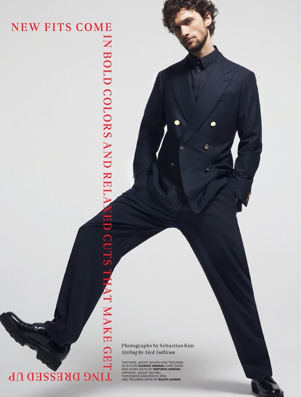 Fashion editorial shot: a slender adult male model in a black double-breasted suit and black lug sole shoes strikes a pose against a neutral backdrop.