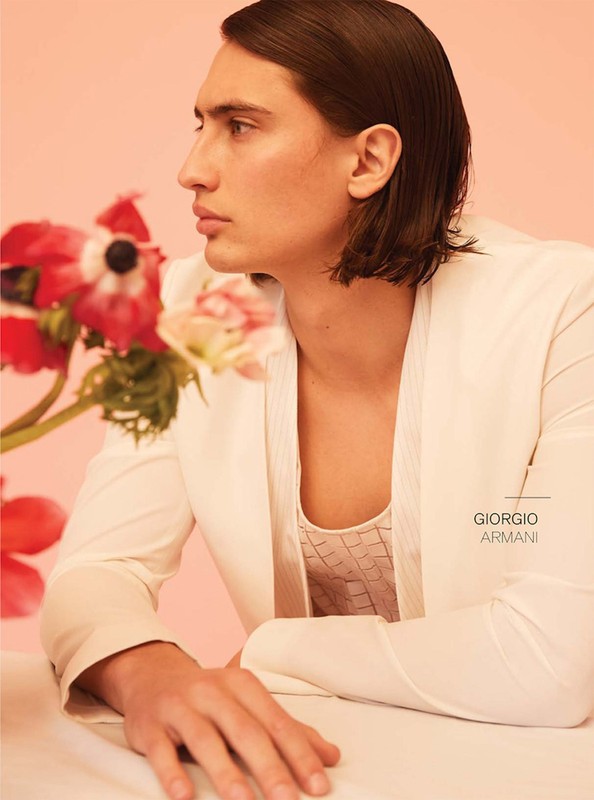 Close-up studio shot of a male model in a white jacket with dark hair, posing against a pink backdrop with flowers. Fashion editorial.