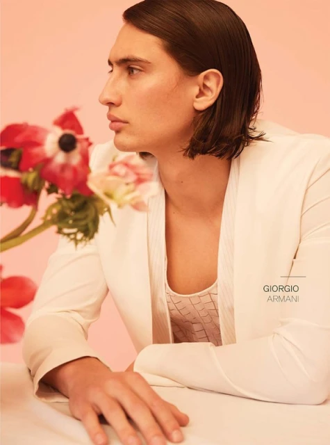 Close-up studio shot of a male model in a white jacket with dark hair, posing against a pink backdrop with flowers. Fashion editorial.