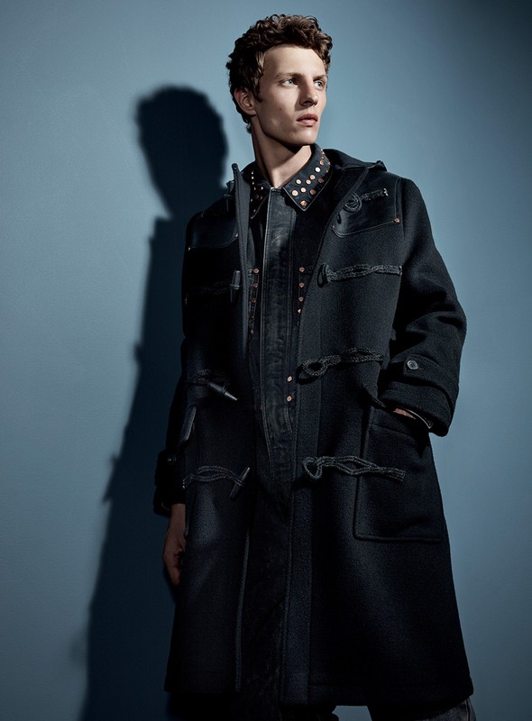 Man in black duffle coat poses in a studio setting, showcasing fashion apparel with a modern and stylish aesthetic against a blue backdrop.