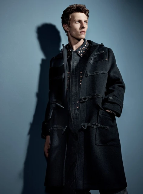 Man in black duffle coat poses in a studio setting, showcasing fashion apparel with a modern and stylish aesthetic against a blue backdrop.