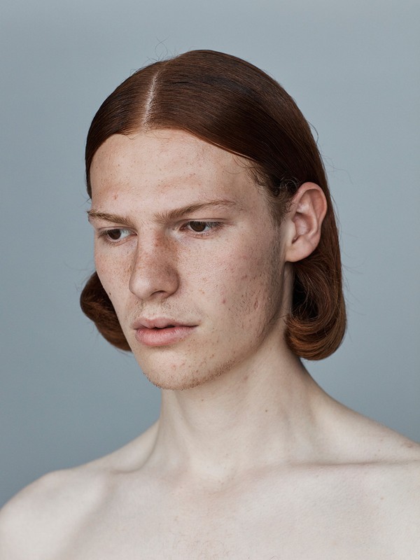 Close-up studio portrait of a redhead adult male with freckles, gazing down with a serious expression, against a neutral grey background.