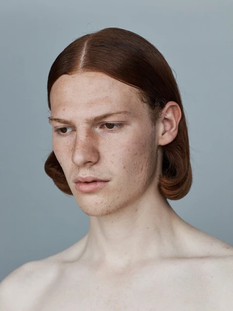 Close-up studio portrait of a redhead adult male with freckles, gazing down with a serious expression, against a neutral grey background.