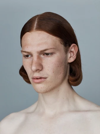 Close-up studio portrait of a redhead adult male with freckles, gazing down with a serious expression, against a neutral grey background.