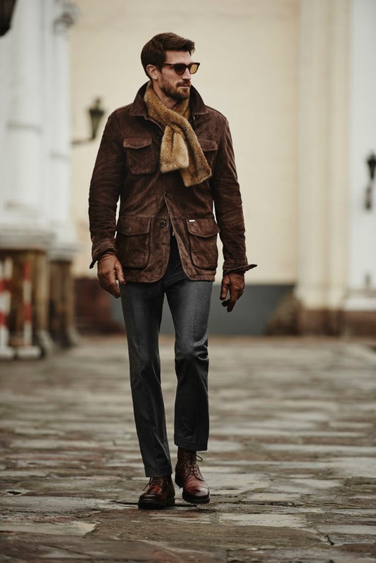 A full shot of a man in a brown jacket, scarf, and jeans walking on a stone path outdoors. He is also wearing sunglasses and brown leather boots.