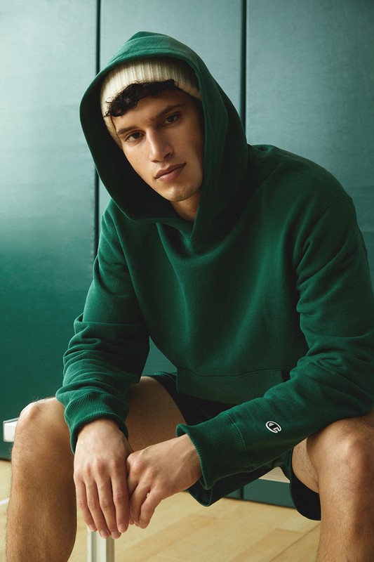 A young adult male sits indoors, wearing a green hooded sweatshirt and a cream-colored beanie, with a neutral expression, against a green background.