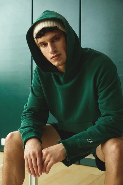 A young adult male sits indoors, wearing a green hooded sweatshirt and a cream-colored beanie, with a neutral expression, against a green background.