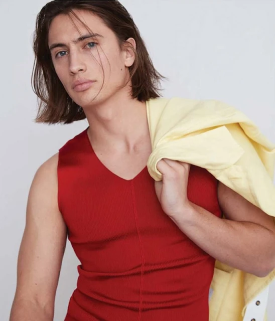 Headshot of a young adult male with long brown hair, wearing a red V-neck shirt and a pale yellow jacket draped over his shoulder.