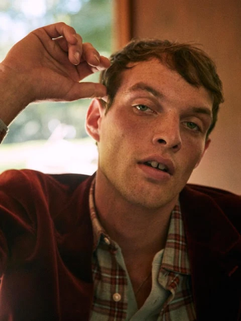 Portrait of a young adult male with brown hair and green eyes, wearing a plaid shirt and red velvet jacket indoors.