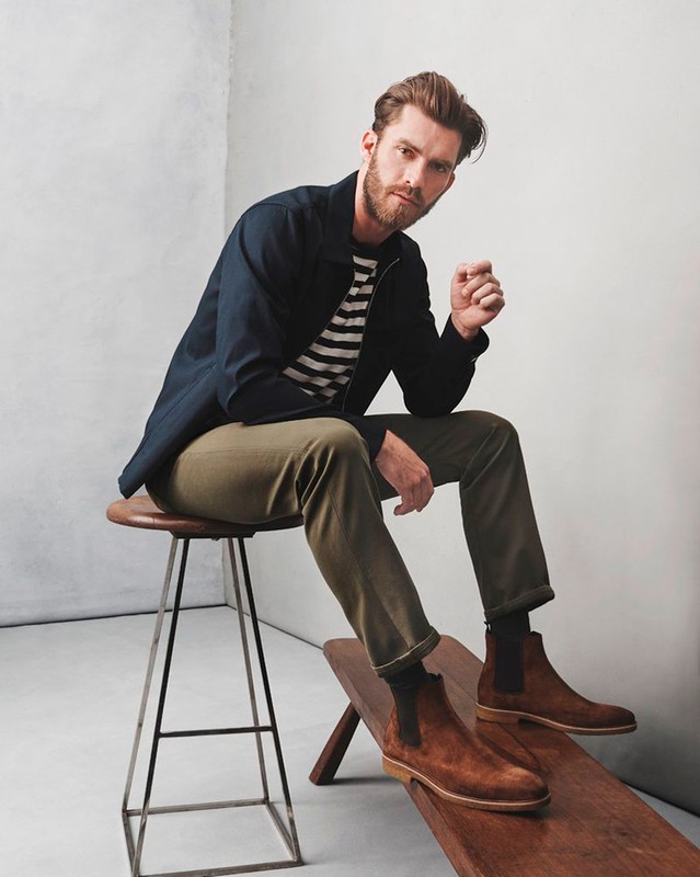 A bearded man sits on a wooden bench in brown boots and a striped shirt, wearing a navy jacket and olive green pants in a studio.