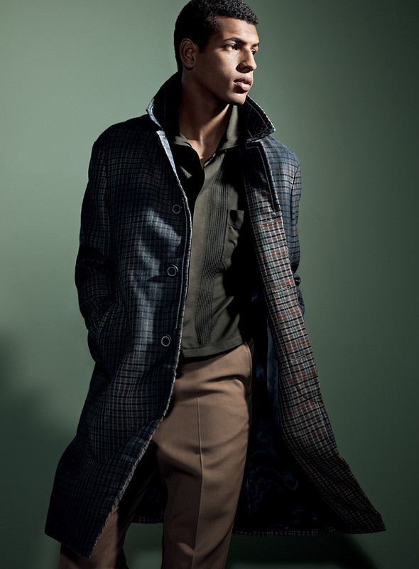 A stylish adult male model showcases a long plaid coat over a polo shirt and slacks in a studio fashion shot on a green background.
