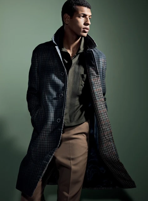 A stylish adult male model showcases a long plaid coat over a polo shirt and slacks in a studio fashion shot on a green background.
