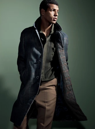 A stylish adult male model showcases a long plaid coat over a polo shirt and slacks in a studio fashion shot on a green background.