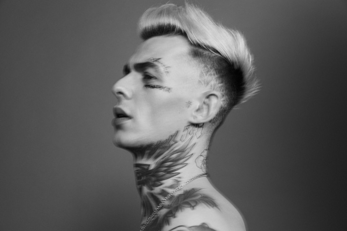 Profile portrait of a tattooed man with styled light hair against a grey backdrop, groomed by James Mooney