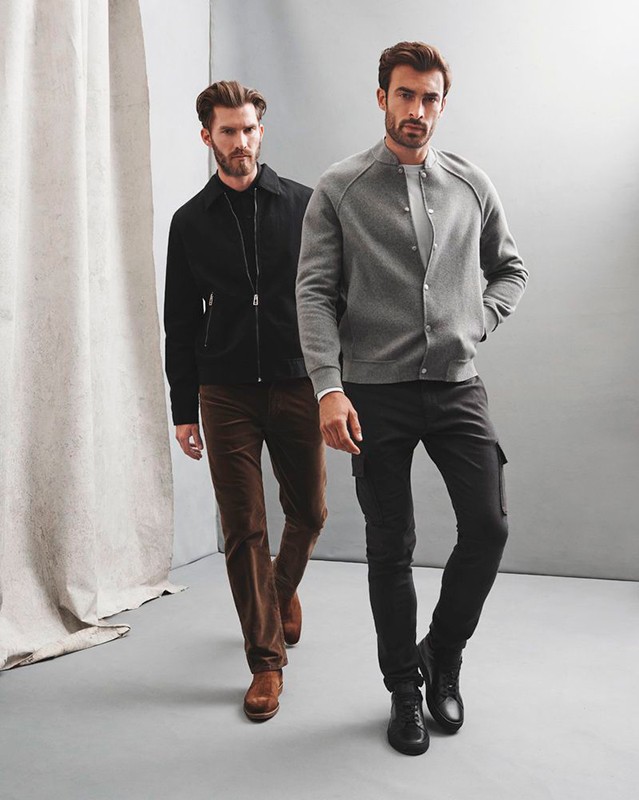 Two adult men in stylish casual wear walking indoors. Man in grey jacket and black pants in front and man in black jacket behind.