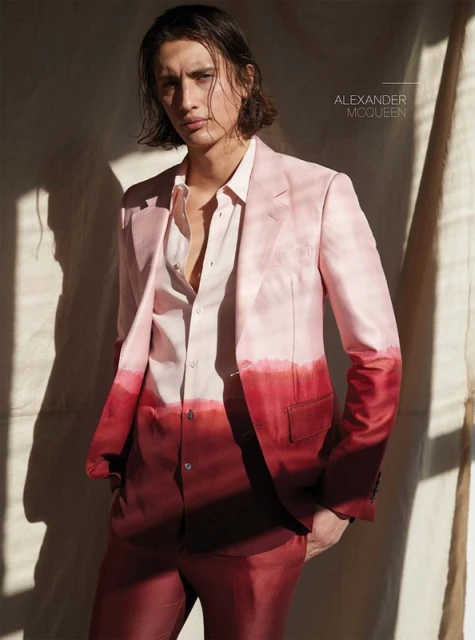 Man poses wearing a pink and red ombre suit, standing in front of a neutral wall with soft lighting creating shadows. Fashion editorial.