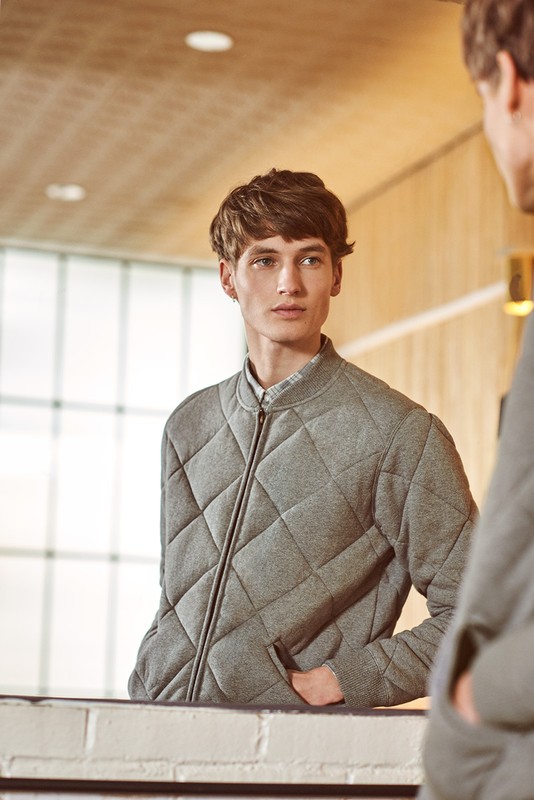 A fair-skinned adult male looks in a mirror while wearing a gray quilted jacket. The background includes a white brick surface.