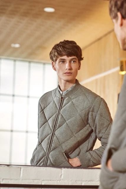 A fair-skinned adult male looks in a mirror while wearing a gray quilted jacket. The background includes a white brick surface.