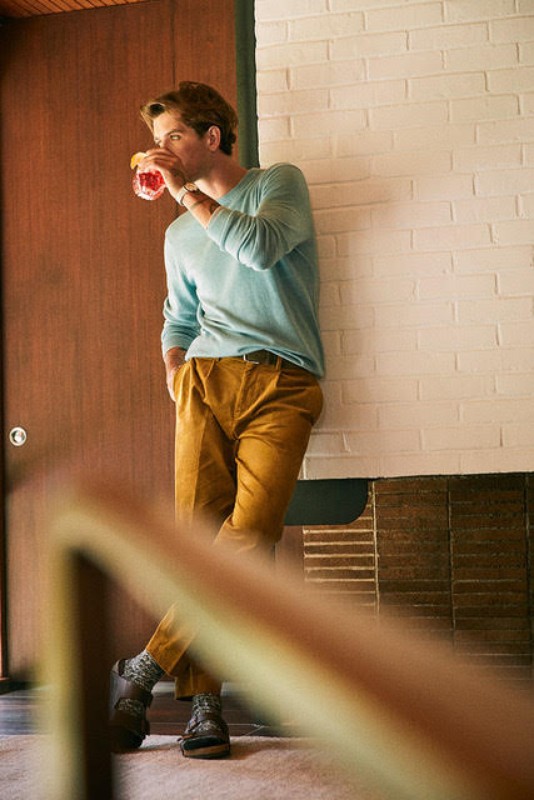 An adult male stands indoors, casually leaning against a white brick wall while drinking a red beverage from a clear glass.