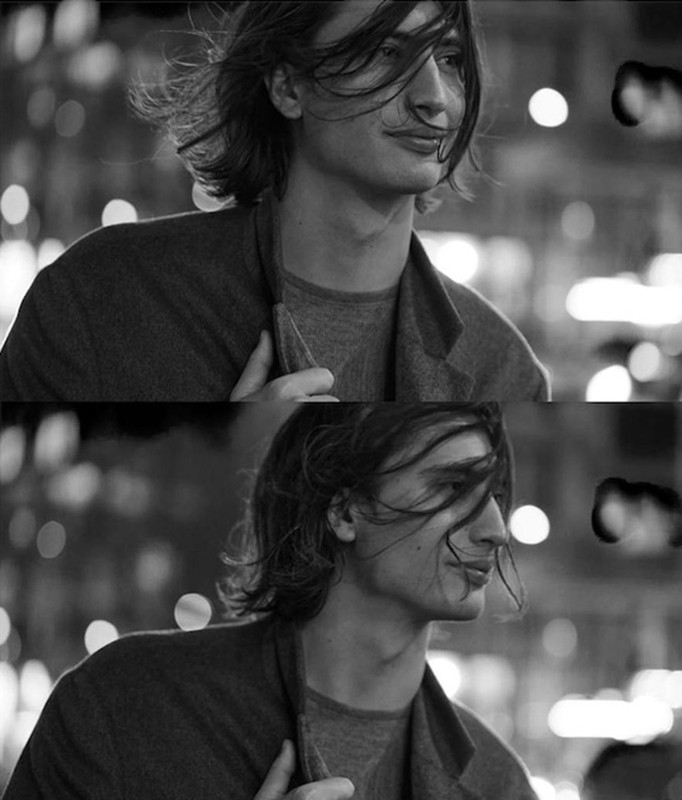 Black and white portrait of a young adult male with shoulder-length hair wearing a jacket in an urban setting with blurred lights.