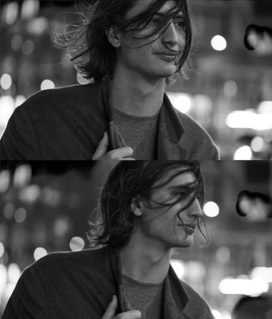 Black and white portrait of a young adult male with shoulder-length hair wearing a jacket in an urban setting with blurred lights.