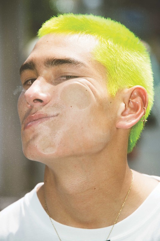 Portrait of a young adult male with green hair pushing his face against glass in an outdoor environment on a bright day.