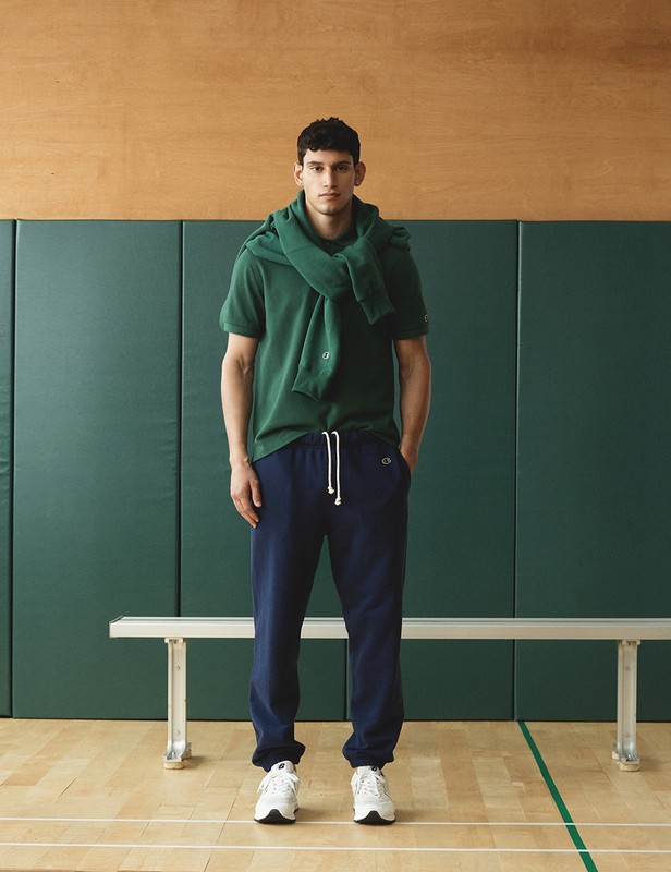 Athletic man in green polo shirt and navy sweatpants in a gymnasium setting, sweater draped over his shoulders, white sneakers.