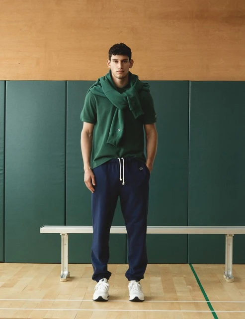 Athletic man in green polo shirt and navy sweatpants in a gymnasium setting, sweater draped over his shoulders, white sneakers.