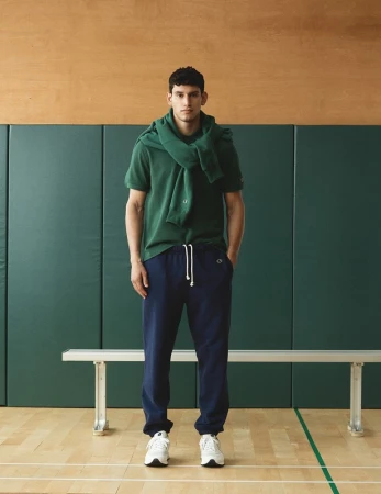 Athletic man in green polo shirt and navy sweatpants in a gymnasium setting, sweater draped over his shoulders, white sneakers.