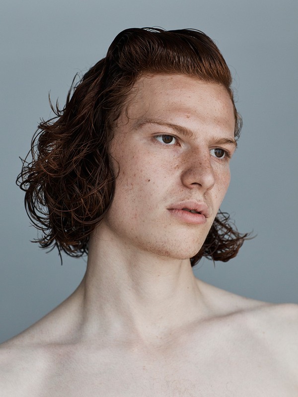 Close-up studio portrait of a young adult male with freckles, reddish-brown hair, and fair skin against a solid grey background, looking right.
