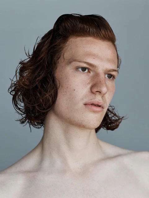Close-up studio portrait of a young adult male with freckles, reddish-brown hair, and fair skin against a solid grey background, looking right.
