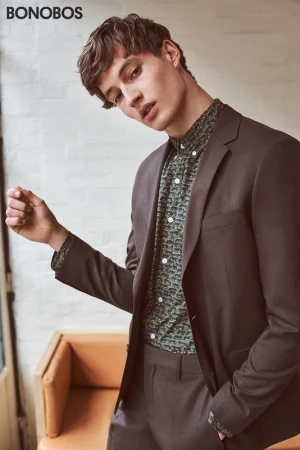 Young man wearing a dark brown suit jacket and patterned green button-down shirt against a brick wall