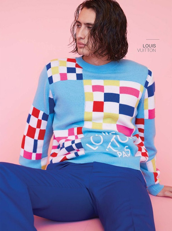 Young male model poses wearing a colorful checkered sweater with blue pants against a pink background in a fashion portrait.