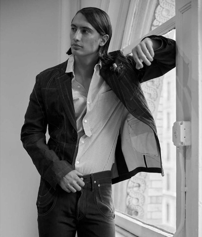 Black and white portrait of a man with long hair wearing a tailored jacket and button-down shirt leaning by a bright window.