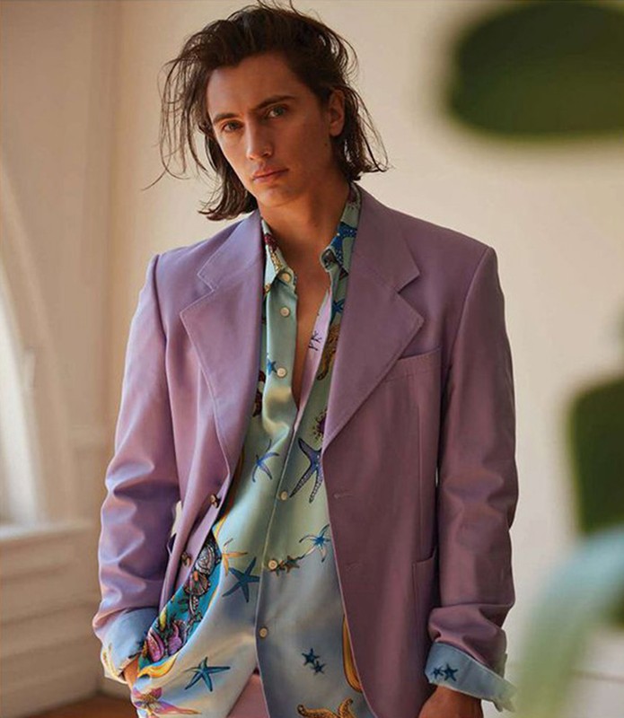 Man in a lilac blazer over a patterned button-down shirt, posing in a photo studio with soft lighting and a blurred background.