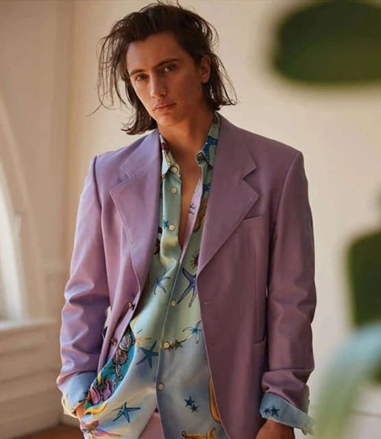 Man in a lilac blazer over a patterned button-down shirt, posing in a photo studio with soft lighting and a blurred background.