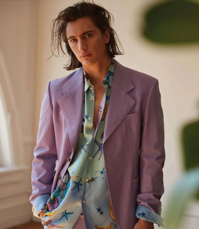 Man in a lilac blazer over a patterned button-down shirt, posing in a photo studio with soft lighting and a blurred background.