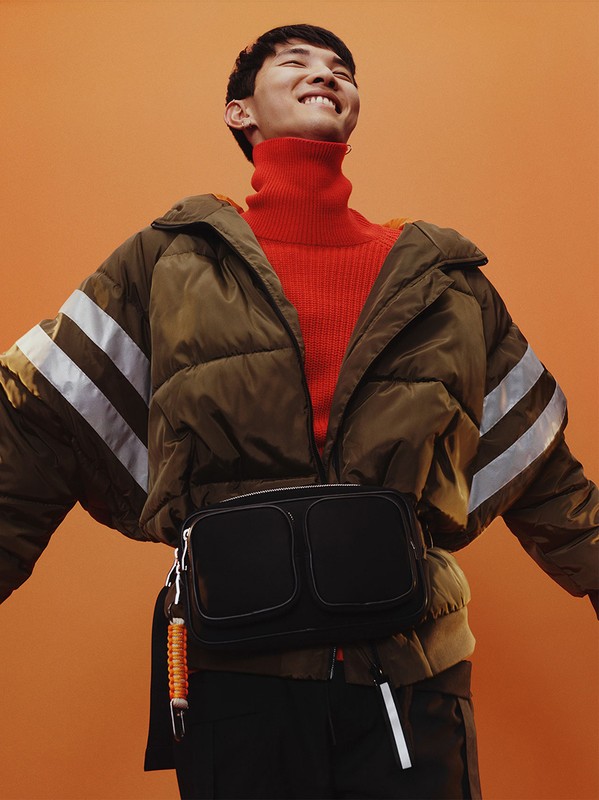 Smiling Asian man poses in a puffer jacket and red turtleneck against an orange backdrop, showcasing a trendy, casual fashion style.